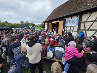 Zahlreiche Erwachsene und Kinder in bunter Herbstkleidung haben sich vor einem großen Fachwerkgebäude in der Alten Schäferei in Ahorn versammelt. Einige sitzen auf Holzbänken, andere stehen. Im geöffneten Scheunentor findet eine Veranstaltung der Kirche Kunterbunt statt. Rechts daneben hängt ein farbenfrohes Banner mit dem Schriftzug „Kirche Kunterbunt Coburg“. Der Himmel ist dicht bewölkt.